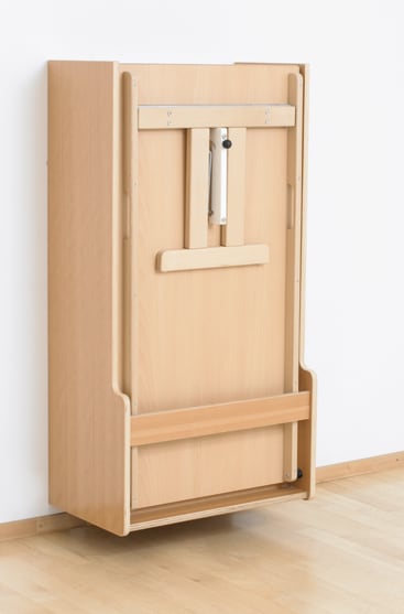 Ein Klapptisch aus hellem Holz, der an einer weißen Wand montiert ist, wird in geschlossener und aufrechter Position gezeigt, wobei Metallscharniere und ein Griff sichtbar sind. Der Boden ist aus hellem Holz.