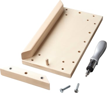 Eine Holzschablone mit einem abnehmbaren Teil, zwei Metallbolzen und einem Schraubendreher mit Bits, dargestellt auf weißem Hintergrund. Die Schablone hat mehrere Löcher und einen Holzpflock zum Ausrichten oder Montieren.