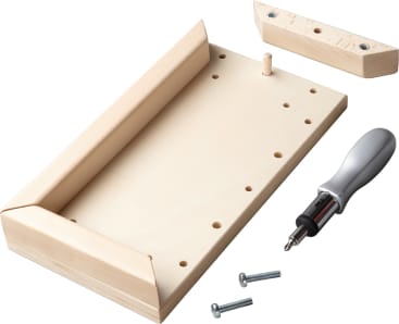 Ein rechteckiges Holzbrett mit abgewinkelten Seitenteilen, einem Holzpflock, mehreren Löchern, zwei Schrauben, einem Schraubenzieher und einem weiteren Holzstück mit zwei Löchern, angeordnet auf einer weißen Fläche.