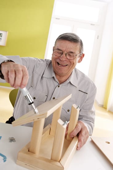 Ein älterer Mann mit Brille lächelt, während er mit einem Schraubenzieher an einem weißen Tisch in einem hellen Raum mit hellgrünen Wänden ein kleines Holzmöbelstück zusammenbaut.