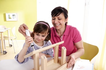 Eine erwachsene Frau und ein junges Mädchen bauen gemeinsam eine Holzkonstruktion zusammen. Das Mädchen benutzt einen Schraubenzieher, während die Frau zusieht und sie anleitet. Sie befinden sich in einem hellen, modernen Raum mit grünen und weißen Wänden.