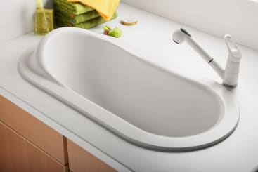 Eine moderne Küche mit einem großen, ovalen weißen Waschbecken, einem weißen ausziehbaren Wasserhahn und hellen Holzschränken. Handtücher, ein Seifenspender und eine kleine Froschfigur stehen auf dem Tresen im Hintergrund.