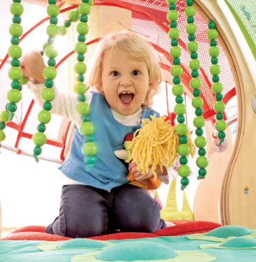 Ein kleines Kind mit blondem Haar kniet auf einer bunten Matte, lächelt aufgeregt und hält eine Puppe in der Hand, die von grünen Perlensträngen umgeben ist, die an einem Spielgerät hängen.