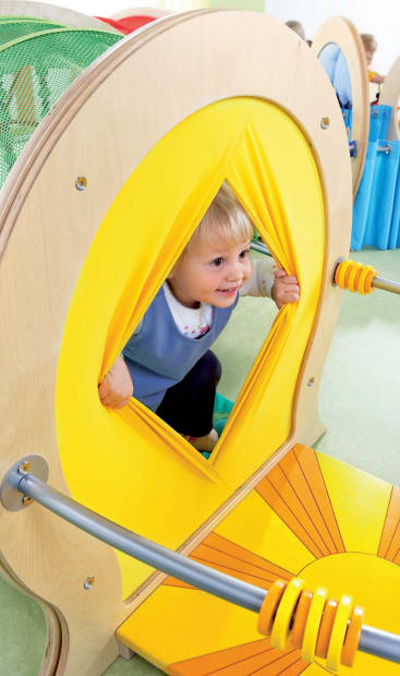 Ein kleines Kind krabbelt durch eine dreieckige gelbe Öffnung in einer hölzernen Spielstruktur. Das Kind lächelt und hält sich an den Seiten des Eingangs fest. Die umliegenden Spielgeräte sind bunt und hell beleuchtet.