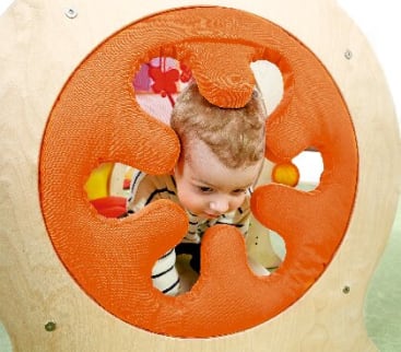 Ein Baby krabbelt durch ein orangefarbenes, strukturiertes, kreisförmiges Spielgerüst mit verschiedenen Ausschnitten und blickt nach unten und vorne. Der Hintergrund zeigt einen Teil eines Spielbereichs.