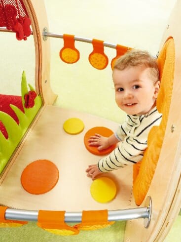 Ein kleines Kind in einem gestreiften Hemd sitzt in einem hölzernen Spielgerät und berührt runde orangefarbene und gelbe Pads auf dem Boden, über denen orangefarbene runde Scheiben hängen.
