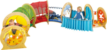 Drei kleine Kinder spielen auf einem Indoor-Klettergerüst aus Holz und Stoff mit Tunneln, Vorhängen und Aktivitätsfeldern in Primärfarben.