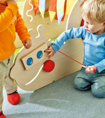 Zwei kleine Kinder spielen mit einem Holzspielzeug mit farbigen Fähnchen, runden Knöpfen und einer roten Schnur. Ein Kind trägt orange, das andere blau, beide sind auf das Spielzeug konzentriert.