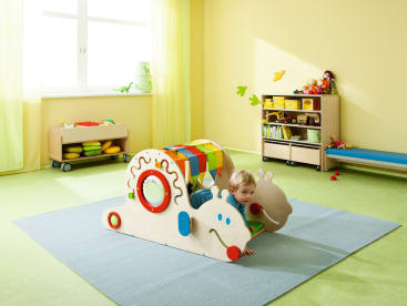 Ein kleines Kind krabbelt durch einen hölzernen Raupentunnel auf einem blauen Teppich in einem hellen, aufgeräumten Spielzimmer mit grünem Boden, gelben Wänden und Regalen mit Spielzeug und Büchern.