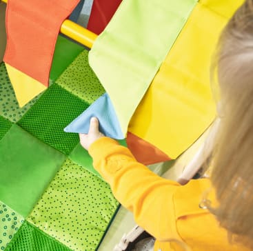 Ein Kind in einem gelben, langärmeligen Hemd greift nach einer weichen, bunten Steppdecke mit grünen, gelben, orangefarbenen und blauen Stoffquadraten, von denen einige eine gepunktete Struktur aufweisen.