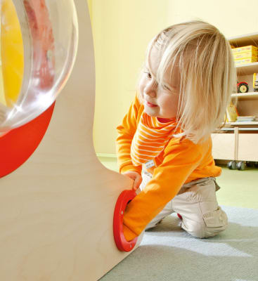Ein kleines Kind mit blonden Haaren und einem orangefarbenen gestreiften Hemd kniet auf dem Boden und dreht einen roten runden Griff an einem hellen Holzspielgerät in einem hellen Raum.