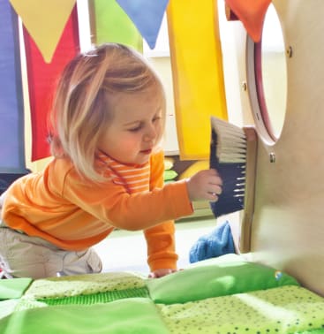 Ein kleines Kind mit blonden Haaren und einem orangefarbenen gestreiften Hemd benutzt eine Bürste, um eine hölzerne Oberfläche in der Nähe eines runden Fensters zu reinigen oder damit zu spielen. Im Hintergrund sind bunte Stoffbanner und weiche grüne Flächen zu sehen.