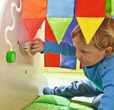 Ein kleines Kind in einem blauen Hemd sitzt auf einer grünen Matte und benutzt einen runden Pinsel auf einer hölzernen sensorischen Spielplatte. Bunte dreieckige Fähnchen hängen über dem Kopf des Kindes.