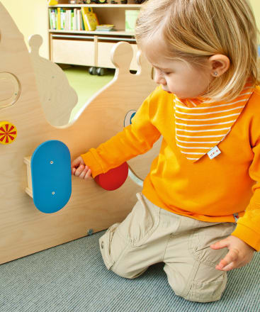 Ein kleines Kind, das ein leuchtend orangefarbenes Hemd und eine beigefarbene Hose trägt, sitzt auf einem Teppichboden und berührt eine blaue Platte auf einer hölzernen Spielstruktur, an der andere farbige Formen befestigt sind. Im Hintergrund sind Regale mit Büchern zu sehen.