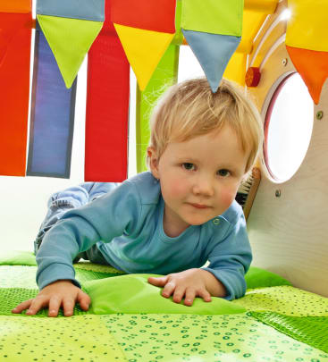 Ein kleines Kind mit blondem Haar und blauem Hemd krabbelt auf einer grün gepolsterten Matte unter bunten Stoffbannern in einem Spielbereich.