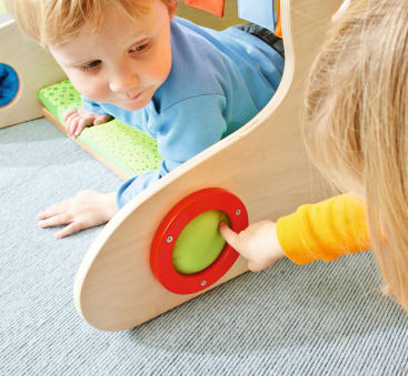 Zwei kleine Kinder spielen zusammen. Ein Kind krabbelt durch eine Holzstruktur, während das andere einen großen, runden, grün-roten Knopf drückt, der an der Seite angebracht ist. Die Szene spielt sich auf einem hellen Teppich ab.