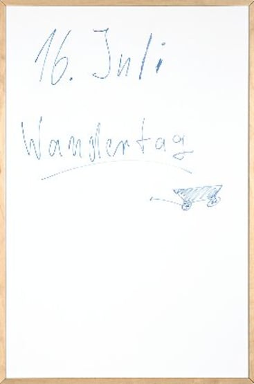Ein Whiteboard mit handgeschriebenem Text in blauer Tinte, auf dem 16. Juli und Wandertag. Unter dem Text ist eine einfache Skizze eines Wagens mit zwei Rädern gezeichnet. Die Tafel hat einen leichten Holzrahmen.