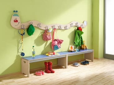 Eine Kindergarderobe in Form einer Raupe ist an einer grünen Wand über einer Holzbank angebracht. An den Haken hängen verschiedene Jacken, Taschen und Hüte, während Schuhe und Stiefel auf dem Boden unter der Bank abgestellt sind.