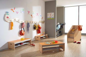 Eine gut beleuchtete Vorschulgarderobe mit Holzbänken, Fächern und Haken für die Taschen und Kleidung der Kinder. An der Wand sind bunte Zeichnungen und eine freundliche Raupe zu sehen. Im Hintergrund sind Kinderwagen und Spielzeug zu sehen.
