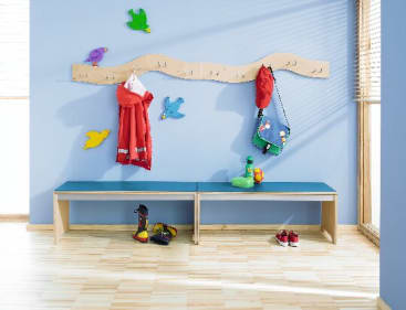Ein Wickelraum für Kinder mit einer blauen Wand, einer Holzbank, vogelförmigen Wandhaken, an denen ein roter Mantel, eine Tasche und ein Hut hängen, sowie Spielzeug und Schuhe auf und unter der Bank. Das Sonnenlicht fällt durch ein Fenster mit Jalousien ein.