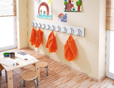 Ein Klassenzimmer mit einem kleinen Holztisch, Hockern, Kunstzubehör und mehreren orangefarbenen Kitteln, die an Wandhaken hängen. Über den Haken hängen Kinderzeichnungen, und durch die Jalousien fällt Sonnenlicht.