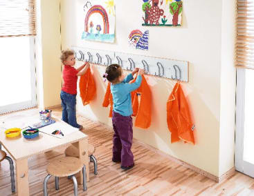Zwei kleine Kinder hängen orangefarbene Kittel an Haken entlang einer Wand in einem Klassenzimmer auf. An der Wand hängen mehrere bunte Kinderzeichnungen. In der Nähe steht ein kleiner Tisch mit Malutensilien und Stühlen.