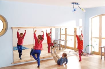 Vier Kinder üben in einem hell erleuchteten Raum vor einem großen Wandspiegel Gleichgewichtsposen. Im Hintergrund sind Hula-Hoop-Reifen und kleine Tische zu sehen.