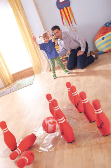 Ein kleines Kind und ein Erwachsener spielen Indoor-Bowling. Das Kind hebt aufgeregt die Arme, als die rot-weiße Kugel mehrere rote Kegel auf einem Hartholzboden umwirft. Der Erwachsene kniet in der Nähe, schaut zu und lächelt.