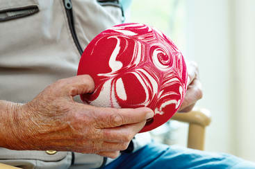 Eine ältere Person hält einen rot-weiß gemusterten Ball, während sie auf einem Stuhl sitzt und die Hände auf dem Schoß ruhen. Das Hemd der Person ist hell gefärbt und teilweise sichtbar.