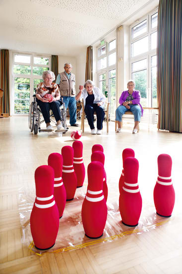 Vier ältere Menschen spielen drinnen eine Partie Bowling mit rosa Kegeln; eine Frau im Rollstuhl rollt eine rosa Kugel auf die Kegel zu, während die anderen zusehen. Große Fenster säumen den Raum und lassen natürliches Licht herein.