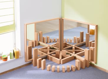 Holzklötze sind in einem Halbkreis auf einem blauen Teppich vor zwei Spiegeln angeordnet, die im rechten Winkel zueinander stehen und symmetrische Reflexionen erzeugen. Der Raum hat grüne Wände und ein Fenster mit Jalousien.