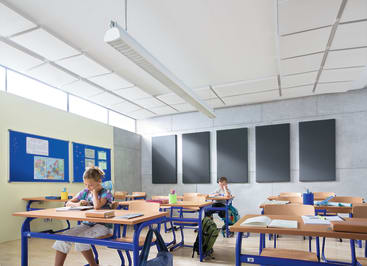 Zwei Kinder sitzen getrennt an Tischen in einem hell erleuchteten Klassenzimmer mit blauen Möbeln, hellen Wänden, Pinnwänden und Schallschutzwänden. Auf oder neben den Tischen stehen Bücher, Wasserflaschen und Rucksäcke.