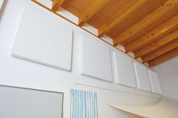 Eine Reihe von fünf großen, weißen, quadratischen Akustikplatten ist hoch an einer weißen Wand unter einer Holzdecke mit sichtbaren Balken angebracht. Darunter sind teilweise eine Tür und eine blau-weiß gestreifte Wanddekoration zu sehen.
