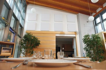 Ein moderner Essbereich mit einem Holztisch, der mit Tellern, Gläsern und Besteck ausgestattet ist. Große Fenster lassen viel natürliches Licht herein. Der Raum hat Holzbalken an der Decke, Zimmerpflanzen und eine Küche, die durch eine offene Tür sichtbar ist.