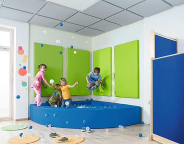 Vier Kinder spielen in einer blauen Ballgrube in einem hell erleuchteten Raum mit grünen Wandpads und schalldichten Deckenplatten. Blaue und weiße Bälle sind verstreut, und es gibt bunte Wanddekorationen und einen Raumteiler.