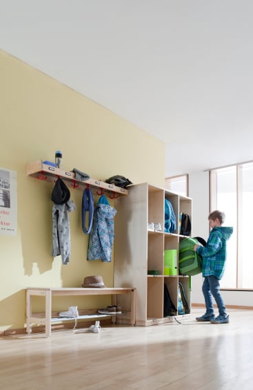 Ein Junge mit einer blau karierten Jacke stellt einen grünen Rucksack in ein Holzfach. Die Fächer befinden sich neben einer Garderobe mit hängenden Jacken, Taschen und Hüten. Auf der Bank und den Regalen darunter befinden sich Schuhe und andere Gegenstände.