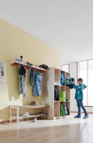Ein kleiner Junge steht in der Nähe von hölzernen Regalfächern und Wandhaken mit Jacken und Taschen in einem hellen, organisierten Eingangsbereich oder in der Garderobe einer Klasse. Er stellt oder holt eine Brotdose aus einem Fach.