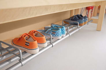 Drei Paar Kinderschuhe - orange, blau mit weißen Punkten und dunkelblau - stehen fein säuberlich auf einem Metallregal unter einer Holzbank. Die Schuhe stehen in einer Reihe auf einem hellen Boden.