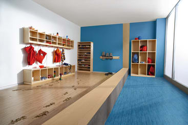 Ein hell erleuchteter, moderner Umkleideraum mit blauen und weißen Wänden, offenen Holzfächern mit roten Uniformen und Ausrüstung, Schuhregalen, Wasserflaschen und Bänken. Auf dem Holzboden sind schmutzige Fußabdrücke zu sehen.