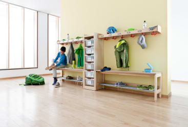 Ein Kind sitzt auf einer Holzbank vor einer Umkleidekabine und zieht sich die Schuhe an. In den Spinden hängen Mäntel, Jacken, Helme und Hüte an Haken, darunter befinden sich Plastikbehälter und auf dem Boden stehen Rucksäcke.