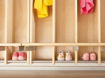 Ein hölzerner Schrank mit drei Paar Kinderschuhen auf dem unteren Regalboden und bunten Kleidungsstücken, darunter gelbe und rosafarbene, die darüber in separaten Fächern hängen.