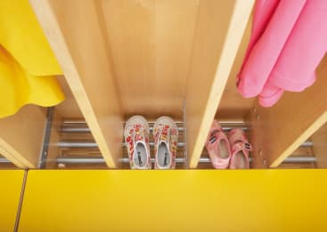 Die Schuhe der Kinder sind in separaten Fächern unter hängenden gelben und rosafarbenen Jacken auf hölzernen Trennwänden untergebracht, davor steht eine gelbe Bank.