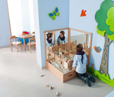 Ein Kind kniet vor einer mit Holzklötzen gefüllten Spiegelecke und baut ein Bauwerk. Im Hintergrund sind bunte Dekorationen, ein Tisch und Stühle zu sehen.