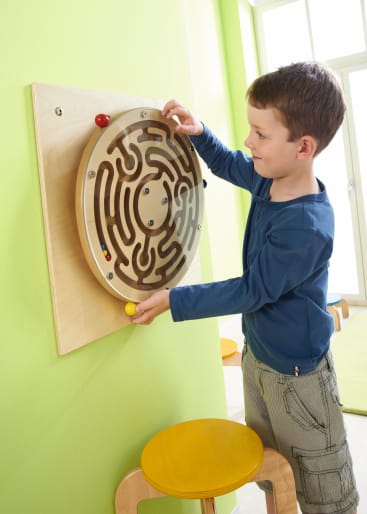 Ein Junge in einem blauen Hemd dreht ein kreisförmiges Holzlabyrinth, das an einer grünen Wand befestigt ist, und führt kleine farbige Kugeln durch die Wege. Ein gelber Hocker steht unter dem Labyrinth. Helles Licht kommt aus einem Fenster im Hintergrund.