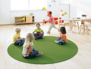 Fünf kleine Kinder sitzen in einem hellen Klassenzimmer. Vier von ihnen sitzen mit gekreuzten Beinen auf einem runden grünen Teppich, während ein Kind in der Nähe läuft. Im Hintergrund sind Regale, Spielzeug und Zeichnungen zu sehen.