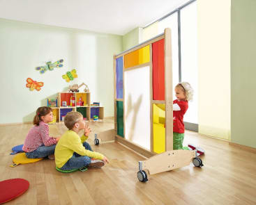Drei kleine Kinder spielen in einem hellen Klassenzimmer. Zwei Kinder sitzen auf Kissen und beobachten, wie ein drittes Kind hinter einem farbenfrohen mobilen Schattenschirm steht und einen Handpuppenschatten wirft. Im Hintergrund sind Regale und Wanddekorationen zu sehen.