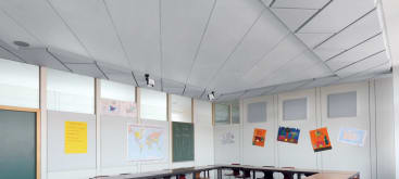 Ein Klassenzimmer mit weißer Akustikdecke, einer grünen Kreidetafel, einer Weltkarte und Kunstwerken der Schüler, die an den Wänden über den Tisch- und Stuhlreihen hängen.