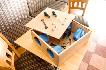 Eine Holzkiste mit kreisrunden Öffnungen und blauem Stoffbezug steht auf einem Tisch. Die Kiste ist teilweise geöffnet und offenbart Schraubenschlüssel, Schraubenzieher, Bolzen und Metallteile im Inneren und auf dem Deckel. In der Nähe stehen zwei Stühle.