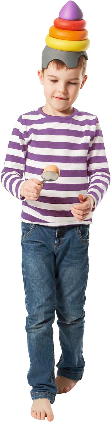 Ein kleiner Junge in blauen Jeans und einem lila-weiß gestreiften Hemd ist barfuß, hält zwei Holzlöffel in der Hand und balanciert bunte, stapelbare Spielzeugringe auf dem Kopf.
