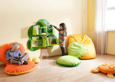 Ein Mädchen in einem gestreiften Hemd greift nach einem Buch in einem grünen, baumförmigen Bücherregal, während ein Junge auf einem orangefarbenen Sitzsack sitzt und liest. Das Zimmer hat gelbe Wände, große Kissen und sonnenbeschienene Holzböden.